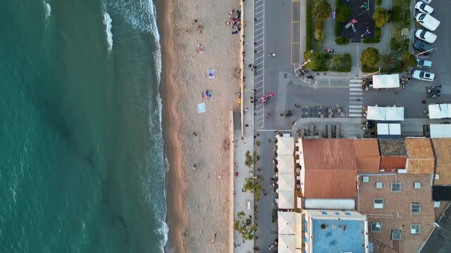 Aerial view of the turquoise sea waves crashing on the sandy beach and people walking along the promenade with palm trees Cefalu, Sicily, Italy.