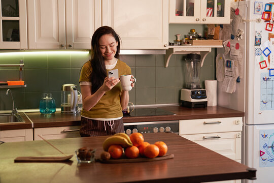 Smiling Woman Holding Cup And Using Phone In Kitchen