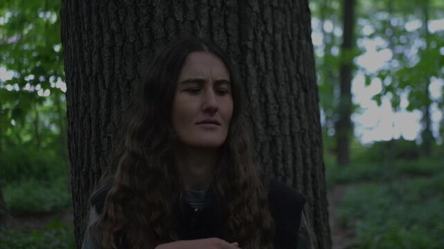 Long-haired woman in a forest swatting mosquitoes near a tree, showing irritation, insect bites, and summer outdoor discomfort.