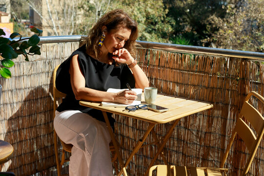 Latina Freelancer Writing in Notebook on Sunny Balcony