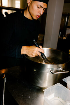 Young chef using a balloon whisk to stir broth in a steel pot