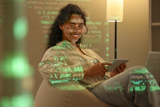 Female African-American programmer with gadgets on beanbag and projection of code in office at night