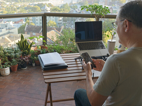 Man Working on a Balcony With Laptop and Phone in a City Setting