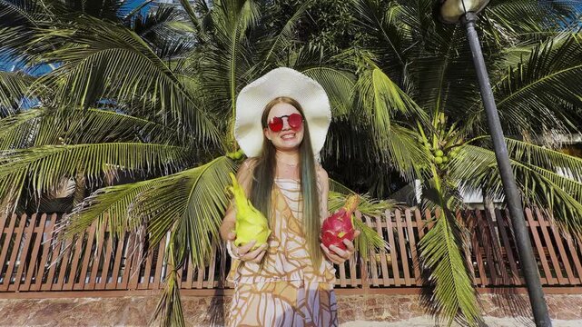 Young woman in tropical setting holding exotic dragon fruits. Summer vacation concept, healthy lifestyle, fresh fruits and bright travel mood under palm trees