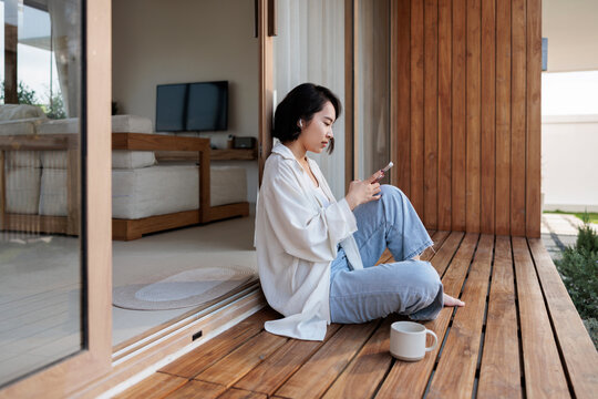 Woman on phone, relaxing on wooden deck with coffee.