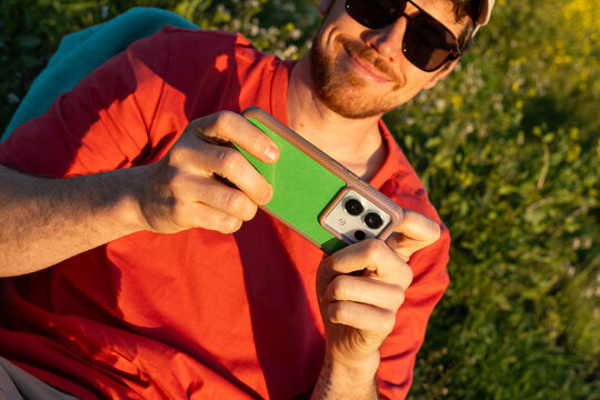 Man Holds Smartphone While Sitting in a Grassy Area During Sunset