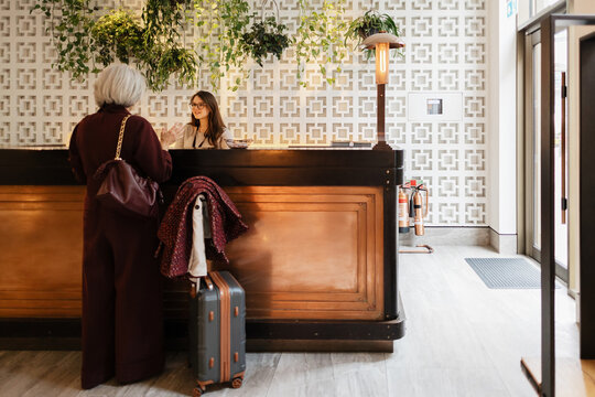 Senior woman booking accommodation at hotel reception with luggage