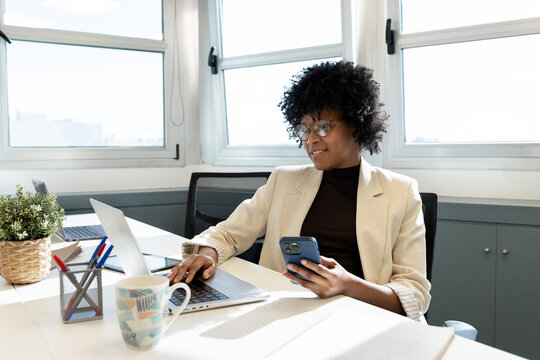 Black businesswoman multitasking with laptop and smartphone at office