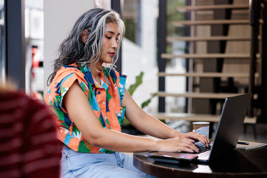 Woman with gray hair works on laptop at cafe.