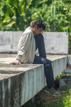 Relaxed man sitting on concrete ledge outdoors under bright sky with greenery