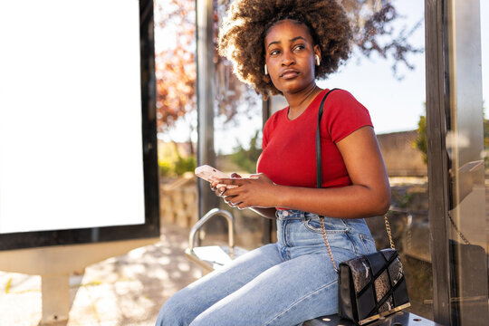 Black woman checking for the arrival of the bus while holding a phone