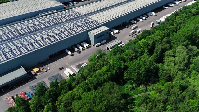 Aerial view of a large industrial warehouse with solar panels on the roof and trucks at loading docks next to a green forest in Coleshill, England, United Kingdom.