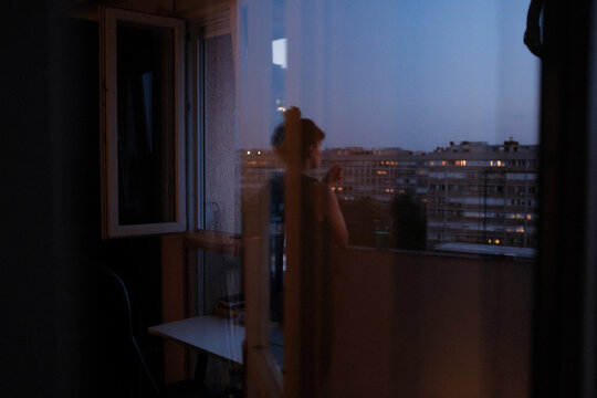 Woman standing by an open window overlooking city at dusk