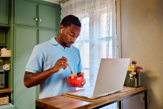 Young Black Man Eating Food while Working on Laptop in Kitchen