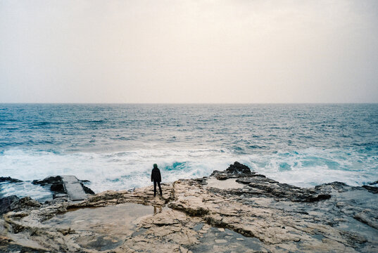 Person standing on rocky shore facing rough sea