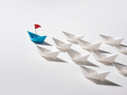 Blue origami boat with red flag leads a fleet of white paper boats