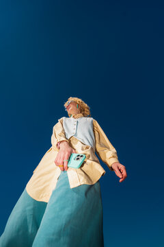 Low angle portrait of woman holding smartphone under blue sky