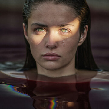 Young woman with tears standing in reflective water under dramatic light and rainbow reflections