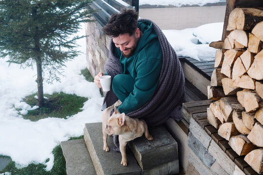 Smiling man with coffee and French bulldog on snowy cabin steps
