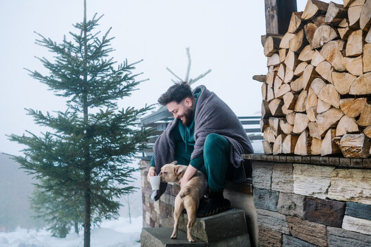 Man petting French bulldog on snowy cabin steps with coffee mug