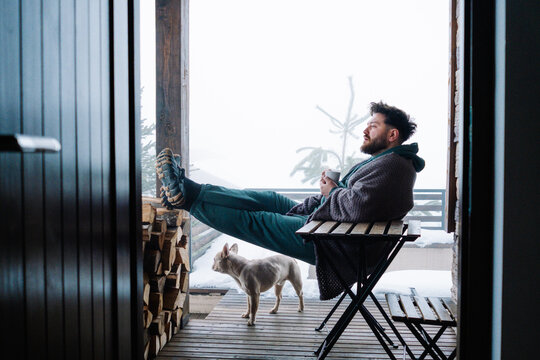 Man relaxing with tea on snowy cabin terrace with French bulldog