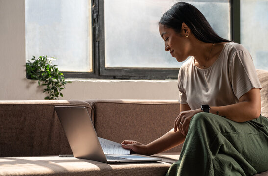 Woman Working On Laptop Looking At Paper Notes, Near Window