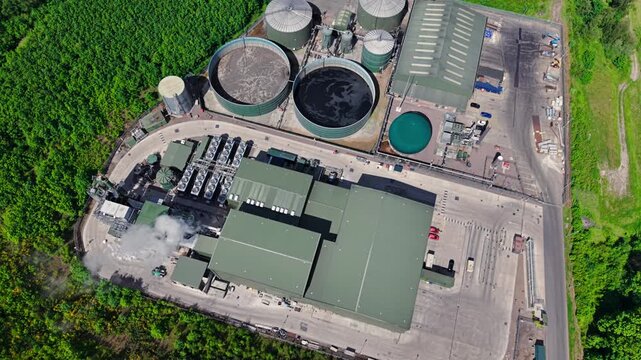 Aerial view of Biogen Atherstone anaerobic digestion plant with circular storage tanks and industrial buildings surrounded by lush green forest in Atherstone, England, United Kingdom.