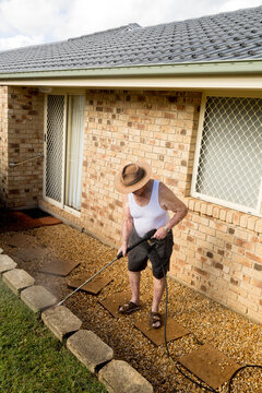 Elderly man pressure washing yard beside brick house
