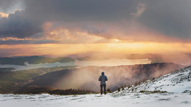 Hiker standing on snowy mountain looking at sunset lake panorama