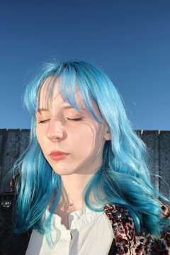 Girl with blue hair stands with eyes closed against blue sky