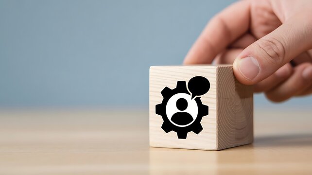 A hand places a wooden cube with a gear and smiley face on a table indoors.