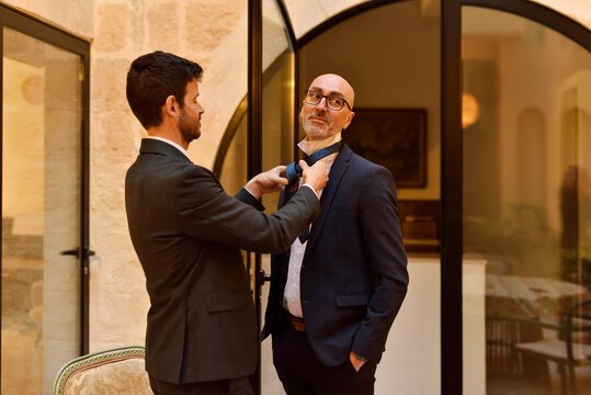 Two men in suits at home adjusting a bow tie before going out