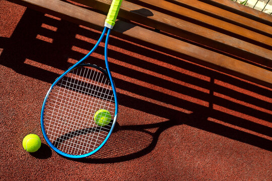 Tennis racket and two balls near a bench on a tennis court.