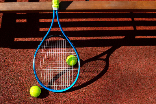 Tennis racket and two balls near a bench on a tennis court.