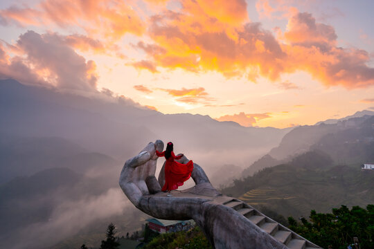 Female tourist in long red dress enjoying the dramatic golden hour view on a giant hand viewpoint in Sapa, Vietnam
