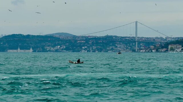 Lonely fisherman in traditional wooden boat in Bosphorus Strait, Istanbul, Turkey. Scenic seascape with flying seagulls flock and majestic suspension bridge on background. Summer travel scene.