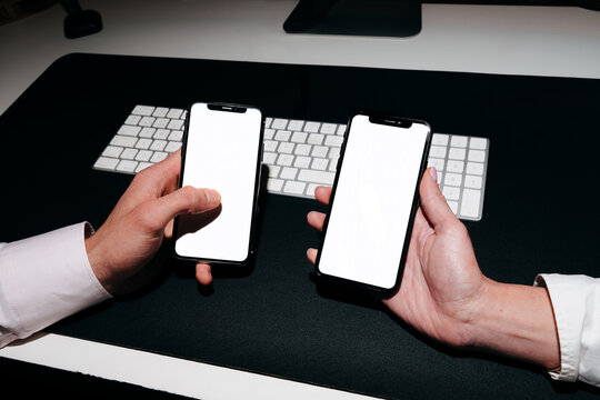 Two Hands Holding Smartphones With White Screen on the Desk