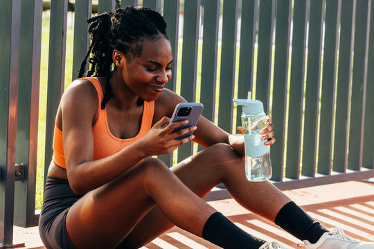 Black woman relaxing after workout with phone and water bottle