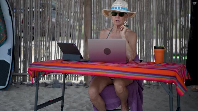 Mature woman has video chat on computer while on a tropical beach while expressing emotions during her vacation.
