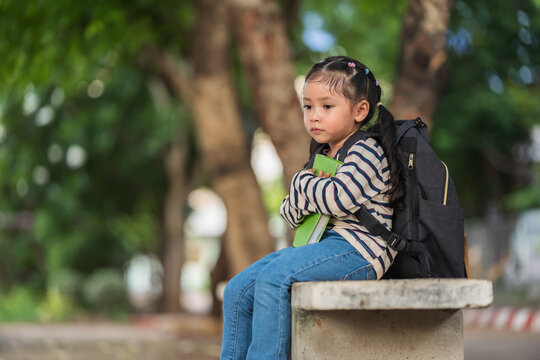 worried child girl sitting on a concrete bench outdoors of school with a book. Education and learning concept.