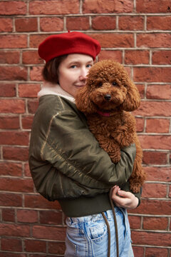 Young woman holds a brown poodle dog against a brick wall 