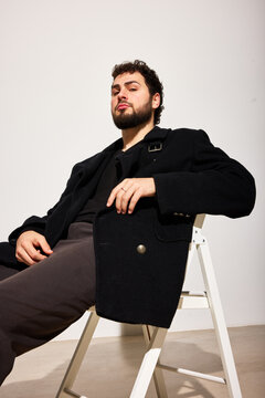 A man with a beard sits casually on a chair in a studio