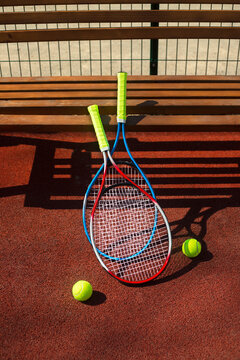 Two tennis rackets and two balls near a bench on a tennis court.