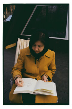 A woman draws in a sketchbook on the street near a house.