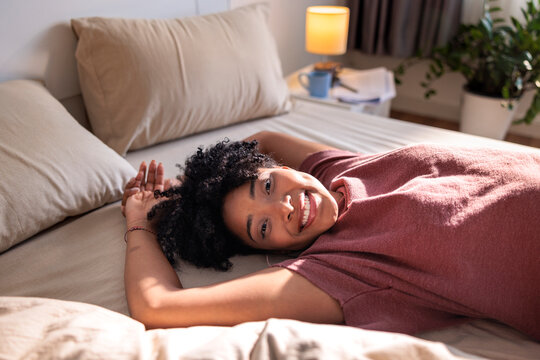 Smiling woman relaxes on bed in sunlit room.