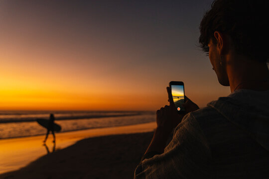 Person capturing sunset moment with smartphone on beach