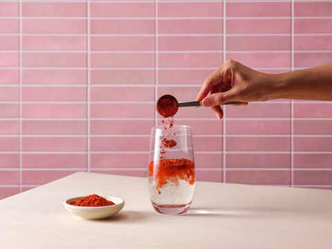 Hand adding vibrant red powder from scoop into clear glass of water
