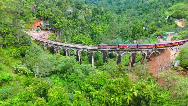 Aerial view of the historic Nine Arch Bridge with a red train crossing through lush tropical forest and tea plantations in Demodara, Uva Province, Sri Lanka.