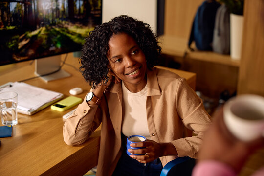 Pleased woman sits at her desk, talking to a colleague