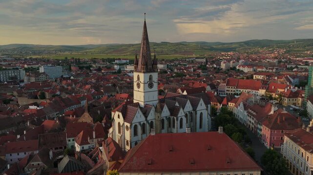 Aerial 4k video of the old town of Sibiu featuring historic red roofs and the tall church tower, with the majestic blue Fagaras mountains visible on the horizon. Sibiu, Transylvania, sunset landscape.